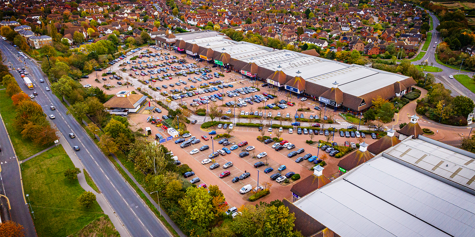 Aerial view of a large retail park with multiple warehouse-style buildings, a spacious parking lot filled with cars, and surrounding roads and greenery. The retail park is situated near a residential area with houses and trees in the background.