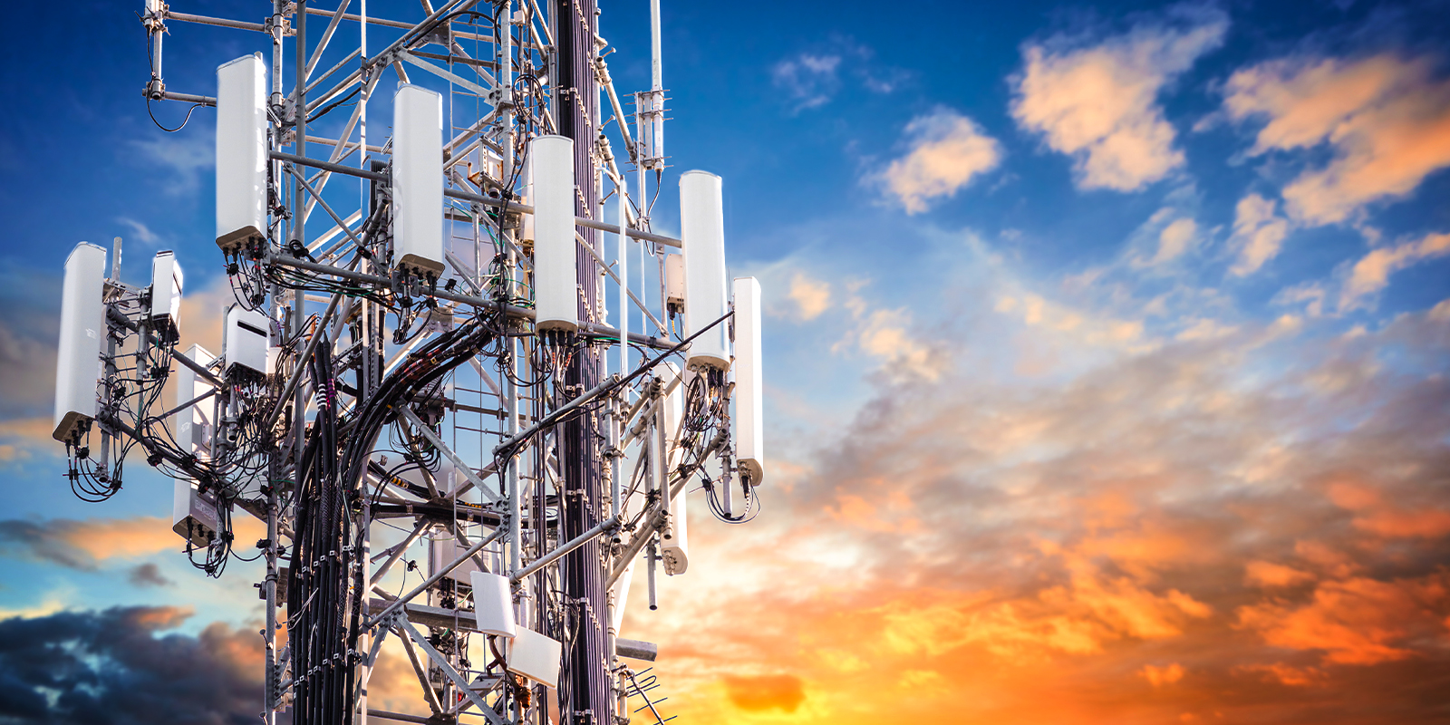 A telecommunications tower equipped with multiple antennas and transmitters, standing against a vibrant sunset sky with scattered clouds. The tower is used for wireless communication and network coverage expansion.