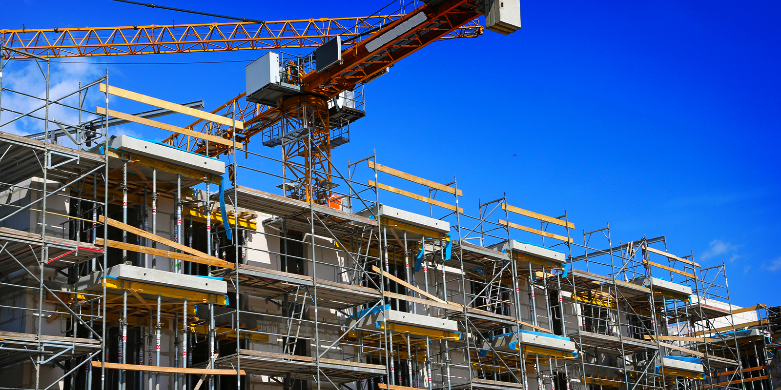 Construction site of a multi-story building with scaffolding, concrete structure, and a large tower crane under a clear blue sky.